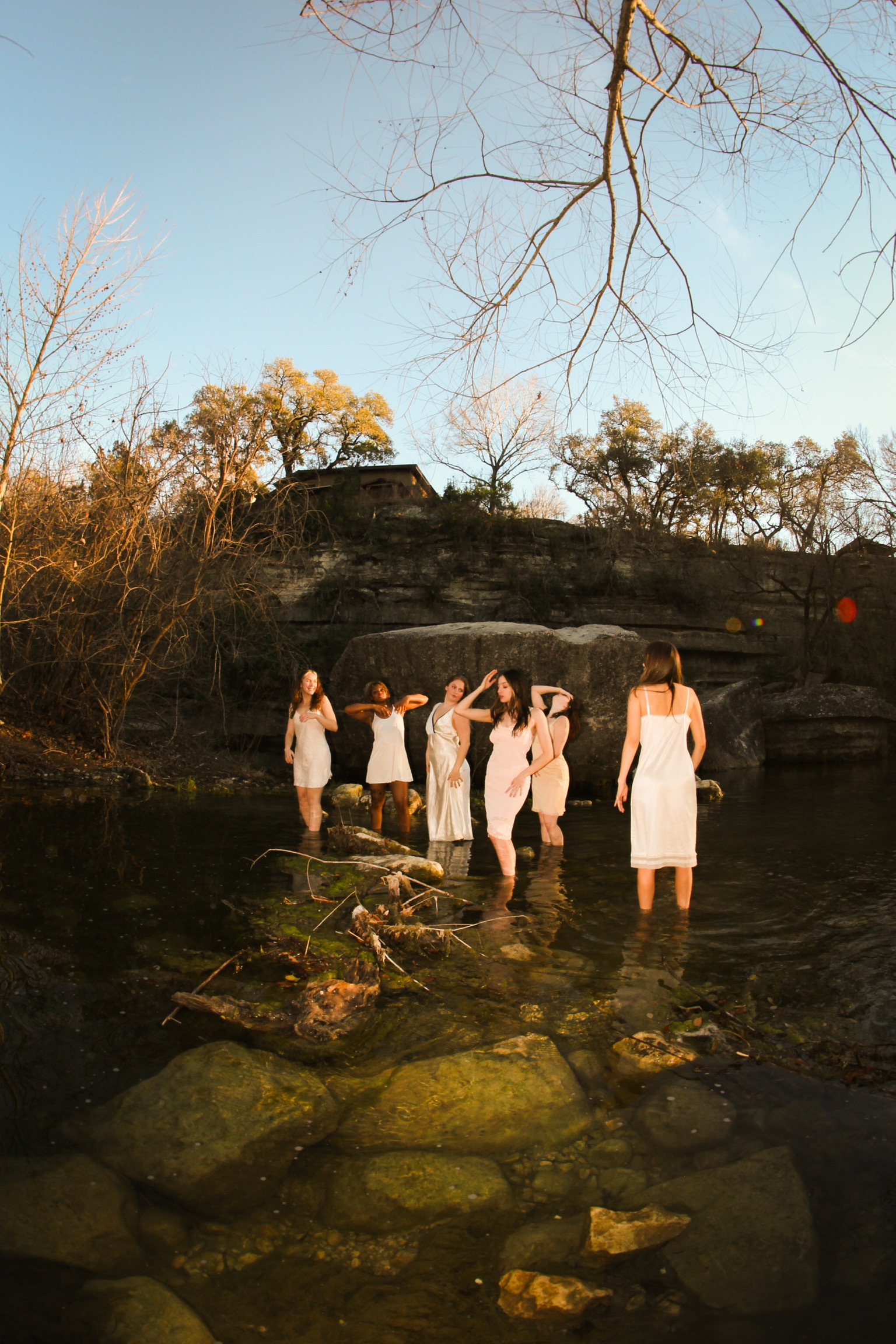 The cast in white slips standing in the creek beneath the limestone bluff at golden hour
